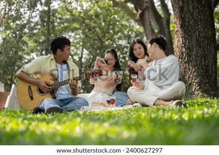 A group of happy young Asian friends is enjoying a picnic in a green park on a summer day together, having fun time, enjoying talking and playing acoustic guitar. Friendship and leisure concepts