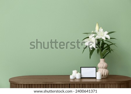 Blank funeral frame, burning candles and vase with lily flowers on wooden cabinet near color wall