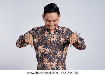 Portrait of smiling young asian man wearing batik shirt pointing index finger down for advertising isolated on white background