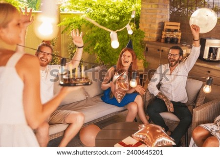 Group of friends having fun at a birthday party, singing while birthday girl is bringing cake