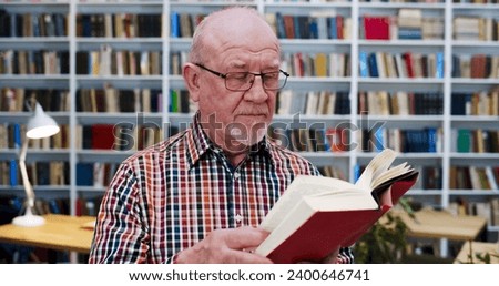 Old Caucasian bald man in glasses and motley shirt reading book in library and studying. Close up of male professor with textbook researching in bibliotheca. Scientific work concept.