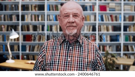 Portrait shot of old Caucasian bald man in motley shirt looking at camera with slight smile in library. Male professor or worker of bibliotheca smiling. Books shelves on background. Senior teacher. Royalty-Free Stock Photo #2400646761