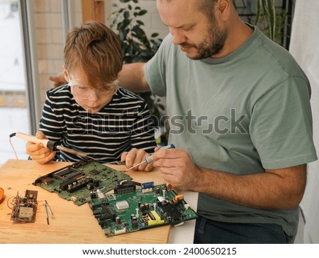 Father with his son spend time together. Close-up of a man teaching his boy at home to solder computer spare parts. Education moment during parenthood. Togetherness concept. Royalty-Free Stock Photo #2400650215