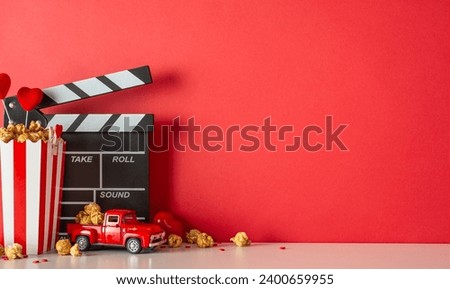 Stay in for a heartwarming movie night on Valentine's Day. Side view picture of a table with a clapperboard, tiny car model, popcorn-filled box, heart decor, and sprinkles against a red wall backdrop