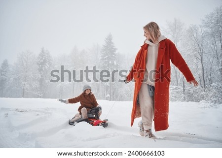 A woman pulls a sled. Mother and her daughter is on the winter meadow and forest. Royalty-Free Stock Photo #2400666103