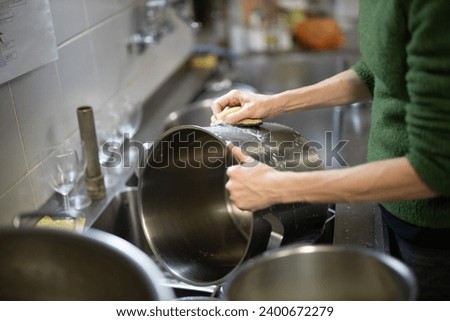 Adult Woman Washes Cooking Pots by Hand in a Restaurant Kitchen Close Up Side View Royalty-Free Stock Photo #2400672279