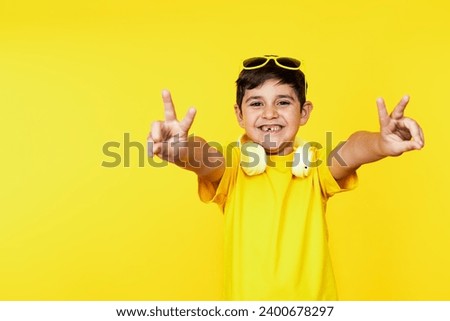 Happy Caucasian kid in yellow tee and headphones makes victory signs, vibrant yellow backdrop