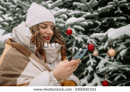 Close up smiling woman using smartphone, standing near Christmas tree, chatting or shopping online, choosing gifts. Girl having fun celebrating New Year on nature. High quality photo