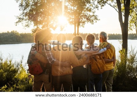 This heartwarming image showcases a group of friends, arms around each other, as they gaze at a lake bathed in the golden light of the setting sun. Their silhouettes against the radiant backdrop speak Royalty-Free Stock Photo #2400688171