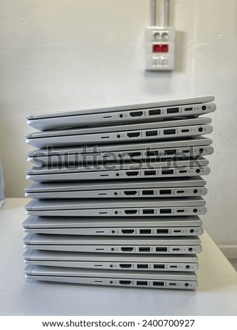 ten silver laptops stacked vertically, one on top of the other.has two USBs, one USB 3.0, one HDMI, a charging port, a mic and a card holder. placed on a gray table and white wall. white and red plugs Royalty-Free Stock Photo #2400700927