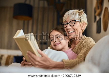 A beautiful grandma and her grandchild, reading a book together, covered in a blanket, lying on the couch. Royalty-Free Stock Photo #2400710829