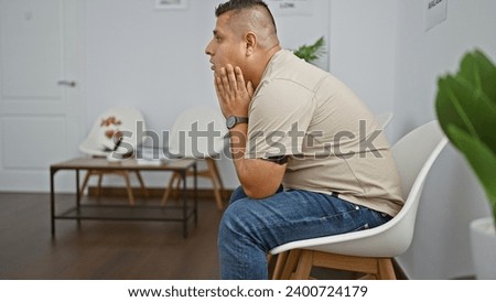 Stressed young latin man alone and frustrated, sitting in a chair, tormented by his problems, exudes an aura of sadness in the sterile indoor waiting room