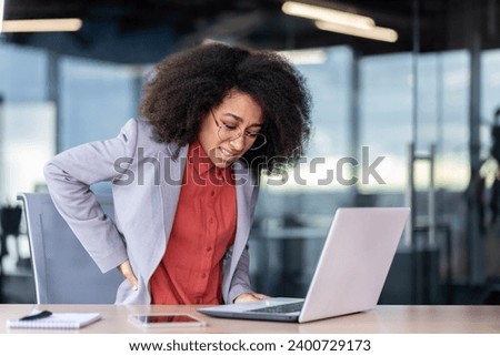Female office worker suffers from back pain, holds her hand on her lower back, posture problems, pinched nerve from sitting work. Royalty-Free Stock Photo #2400729173