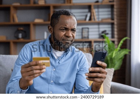 Upset and sad man sitting at home on couch in living room, cheated senior mature african american man displeased holding phone and bank credit card, rejected fund transfer error, account block. Royalty-Free Stock Photo #2400729189
