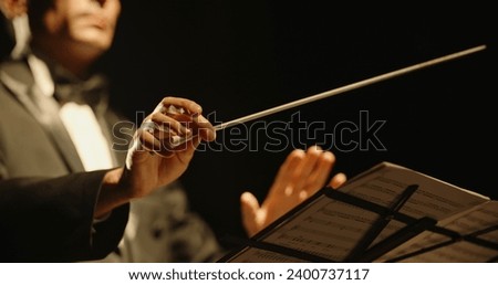 Male orchestra conductor controlling music in orchestra pit by movement of his hands and white baton, studio shot on black background  Royalty-Free Stock Photo #2400737117