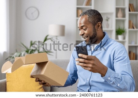 Happy and satisfied buyer received cardboard box in mail, satisfied with speed of delivery of a gift for an online store, an African American man with a phone in hands confirms receipt of the goods. Royalty-Free Stock Photo #2400761123