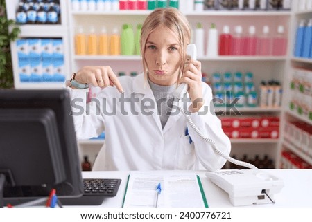 Young caucasian woman working at pharmacy drugstore speaking on the telephone pointing down looking sad and upset, indicating direction with fingers, unhappy and depressed.  Royalty-Free Stock Photo #2400776721