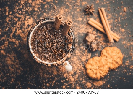 Homemade spicy hot chocolate drink with cinnamon stick, star anise, grated chocolate in enamel mug on dark background with cookies, cacao powder and chocolate pieces, top view.