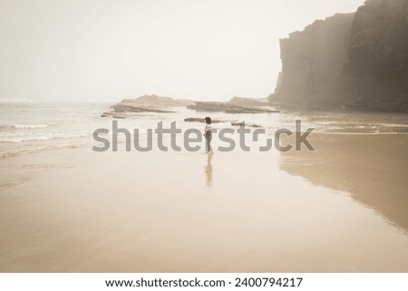 A little girl wearing a dress stands on the sand overlooking and looks over the see in a misty day in Playa de las Catedrales (Cathedrals Beach) in Ribadeo, Galicia, Spain Royalty-Free Stock Photo #2400794217