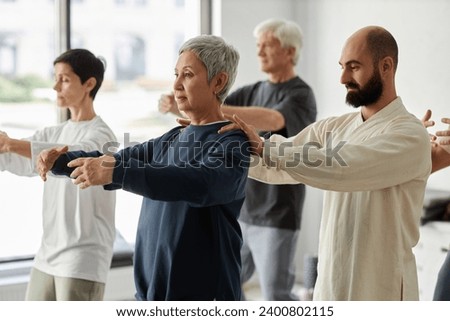 Side view of bearded male trainer correcting shoulder position of gray-haired senior woman during qigong exercise Royalty-Free Stock Photo #2400802115