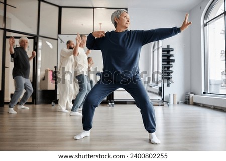 Full shot of gray-haired senior woman dressed in blue sweatsuit practicing qigong archer pose in gym while looking away Royalty-Free Stock Photo #2400802255