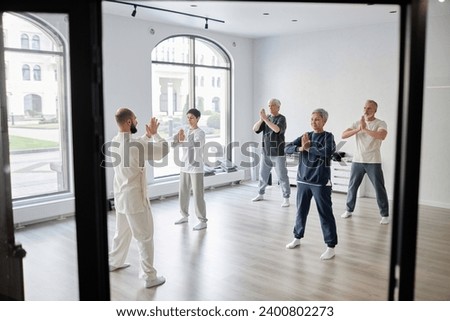 Wide angle shot of mixed group of senior qigong students repeating prayer pose exercise after male trainer in spacious light gym room Royalty-Free Stock Photo #2400802273