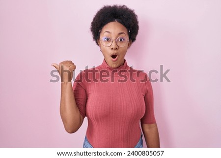 Beautiful african woman with curly hair standing over pink background surprised pointing with hand finger to the side, open mouth amazed expression. 