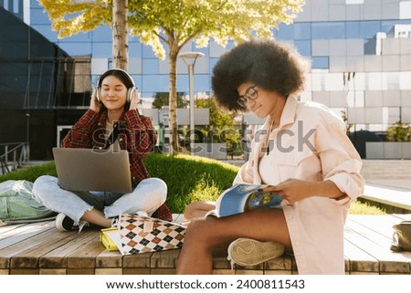 Two smiling multinational female students with laptop studying together while sitting on bench outdoors