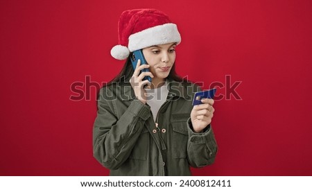 Young beautiful hispanic woman arguing by smartphone holding credit card celebrating christmas hat over isolated red background