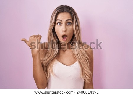 Young blonde woman standing over pink background surprised pointing with hand finger to the side, open mouth amazed expression. 