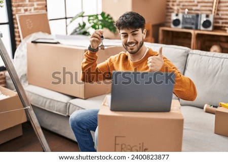 Hispanic man with beard holding keys of new home doing video call with laptop smiling happy and positive, thumb up doing excellent and approval sign 