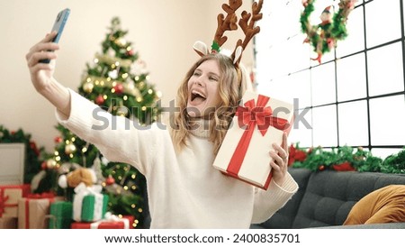 Young blonde woman making selfie by smartphone holding christmas gift at home