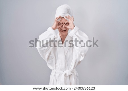 Blonde caucasian woman wearing bathrobe doing ok gesture like binoculars sticking tongue out, eyes looking through fingers. crazy expression. 