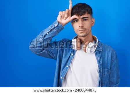 Young hispanic man standing over blue background making fun of people with fingers on forehead doing loser gesture mocking and insulting. 