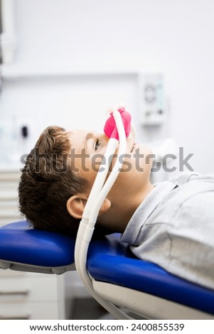 Fear of the dentist! Vertical photo of a little boy sits in a dentist's office wearing a nasal mask breathing nitrous oxide to relax. Concept of feeling relaxed with laughing gas. Royalty-Free Stock Photo #2400855539