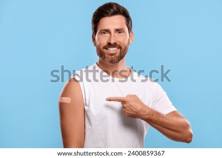 Man pointing at sticking plaster after vaccination on his arm against light blue background Royalty-Free Stock Photo #2400859367
