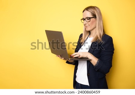 Smiling cheerful funny beautiful attractive young woman 40s wearing basic casual jacket standing working on laptop pc computer isolated on yellow colour background studio portrait