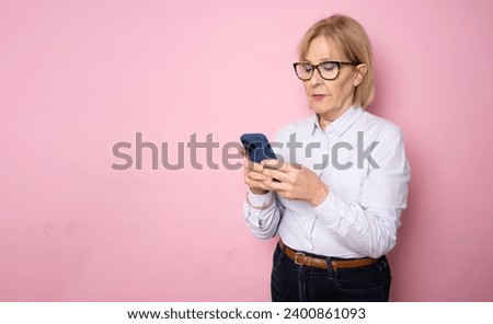 Happy mature senior woman holding smartphone using mobile online apps, smiling old middle aged lady texting sms message chatting on phone looking at cellphone isolated on pink studio background