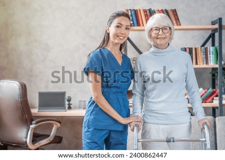 Portrait of senior woman and supporting nurse at home looking at the camera. Rehabilitation process, moving disability, fractures and traumas, injuries. Old woman and doctor image Royalty-Free Stock Photo #2400862447