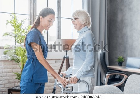 Female asian nurse helping senior woman to get up with walker at home, rehabilitation after injuries, traumas. Pain in muscles, fractures of senior old elderly patients Royalty-Free Stock Photo #2400862461