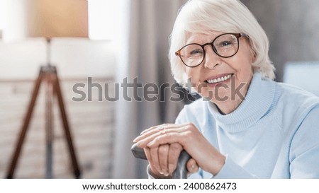 Portrait of happy mature woman in eyeglasses holding cane while sitting on sofa at home looking at camera. Active seniors concept. Lonely grandmother with walking moving disability Royalty-Free Stock Photo #2400862473