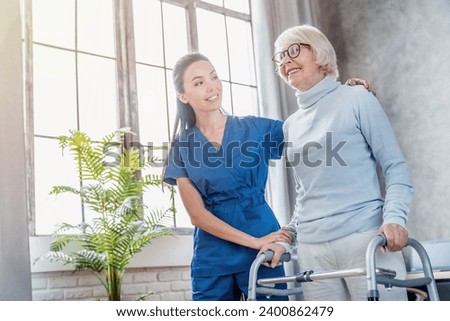 Female asian nurse helping senior woman to walk with walker at home. Rehabilitation after injuries, traumas, walking disability and movement impairment. Senior life concept Royalty-Free Stock Photo #2400862479