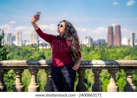 Smiling young Latin woman tourist with long hair in casual clothes and sunglasses taking photo with smartphone in Chapultepec Castle in Mexico city against blurred cityscape background of Reforma stre