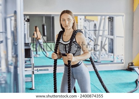 Portrait young strong sporty fit girl in sports clothes training or workout with battle ropes at gym. Woman posing with ropes. Royalty-Free Stock Photo #2400874355