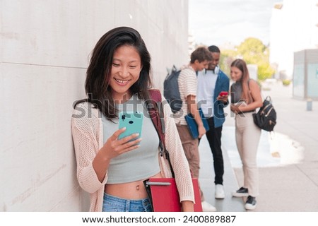 Joyful college student lady watching social media content in her smartphone while a group of colleagues having fun with a cellphone standing outside at university campus. Young woman chatting
