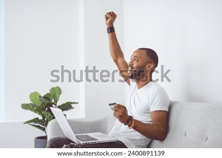A Young happy man on laptop with credit card on couch in light livingroom