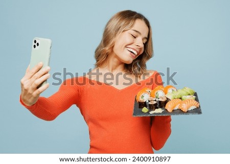 Young happy woman wear orange casual clothes doing selfie shot on mobile cell phone hold eat raw fresh sushi roll served on black plate Japanese food isolated on plain blue background studio portrait