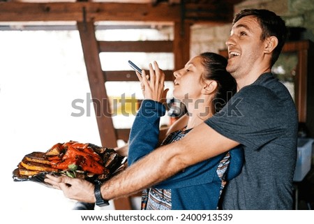 Young woman takes a picture on a smartphone of a dish with baked vegetables in the hands of a laughing man