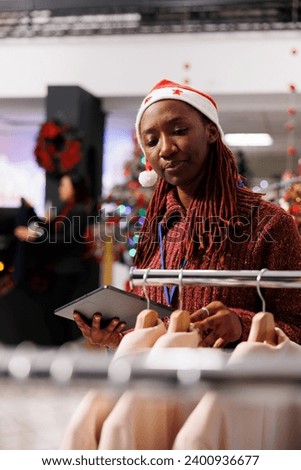 Sales manager checks inventory list with clothing items in retail store, working on stock during christmas holiday season. Woman with santa hat counting merchandise on racks, festive boutique.