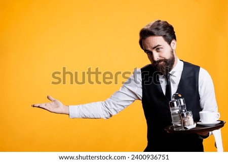 Friendly gracious waiter makes way for people to enter restaurant, providing great service and amazing food at five star place. Young adult with suit and tie posing against yellow background. Royalty-Free Stock Photo #2400936751